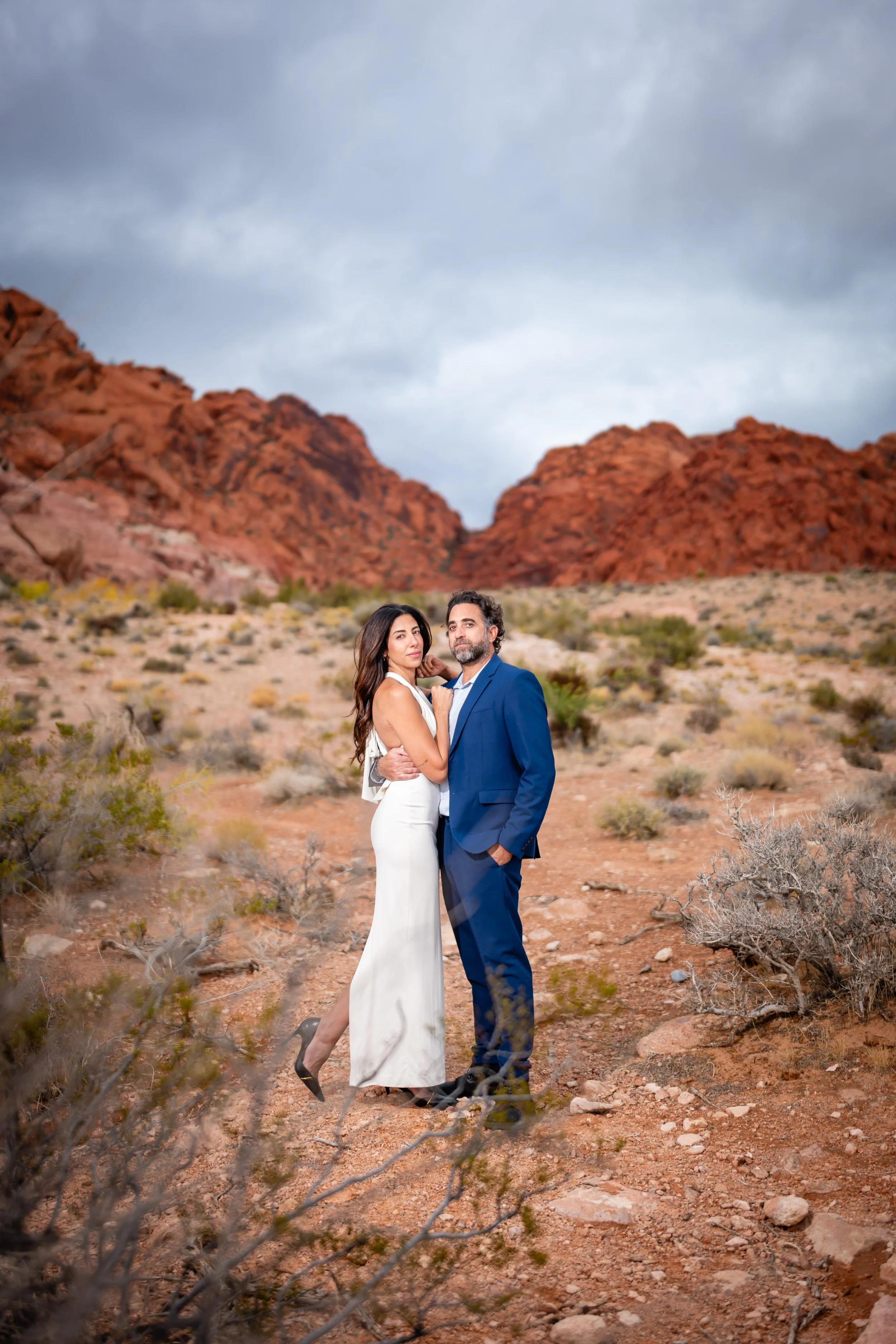 A couple dressed in formal attire standing in a desert landscape with red rock formations in the background, under a cloudy sky.
