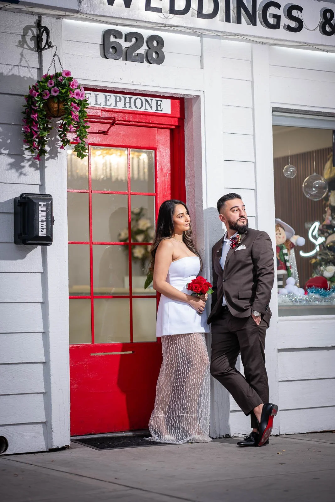 A couple dressed in formal attire standing outside a wedding and event venue with a red door, white exterior, and holiday decorations, including a Christmas tree and whimsical characters inside the window. The woman is holding a bouquet of red roses,