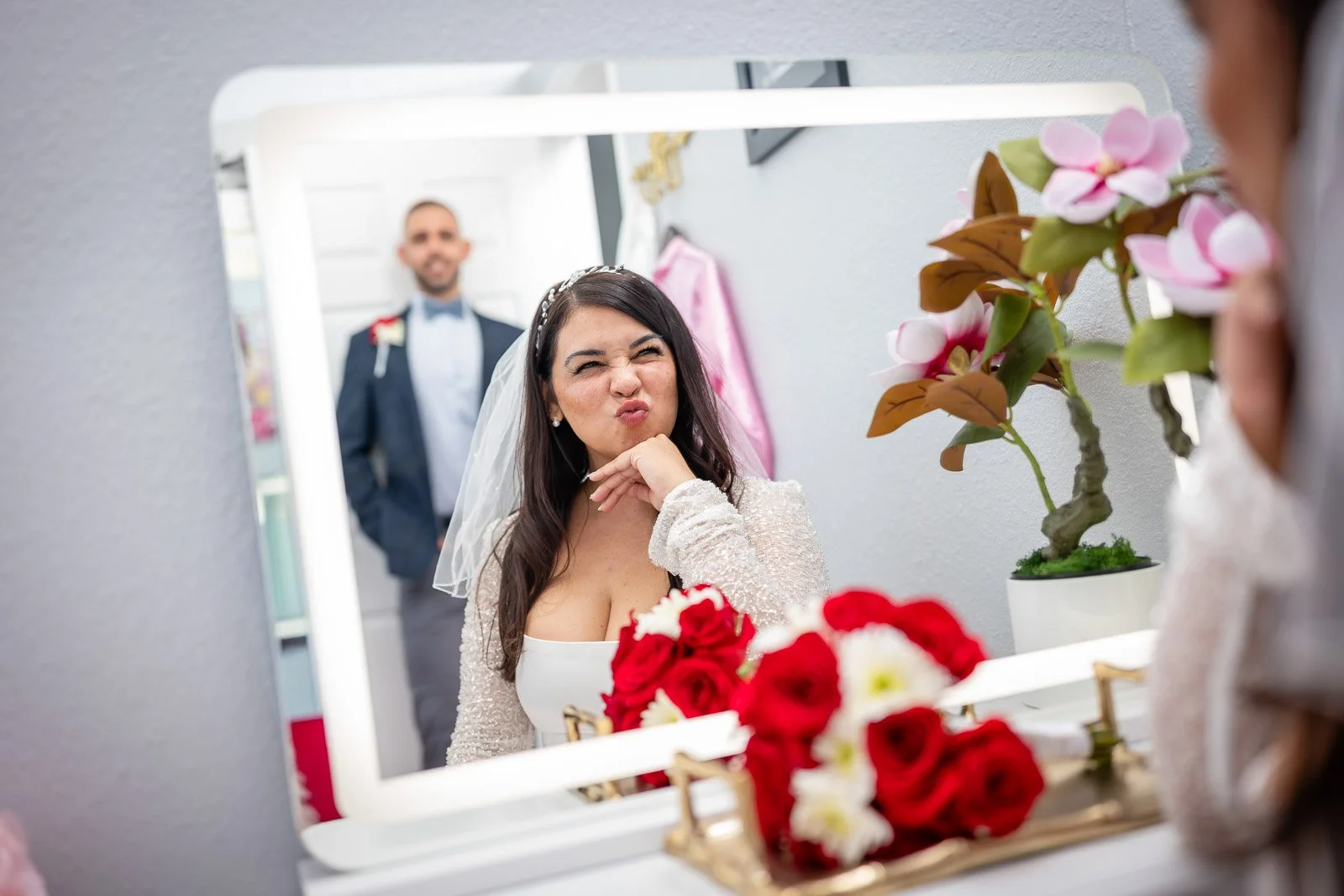 Bride making a funny face while looking in a mirror, with a groom standing in the background, decorated for wedding day.