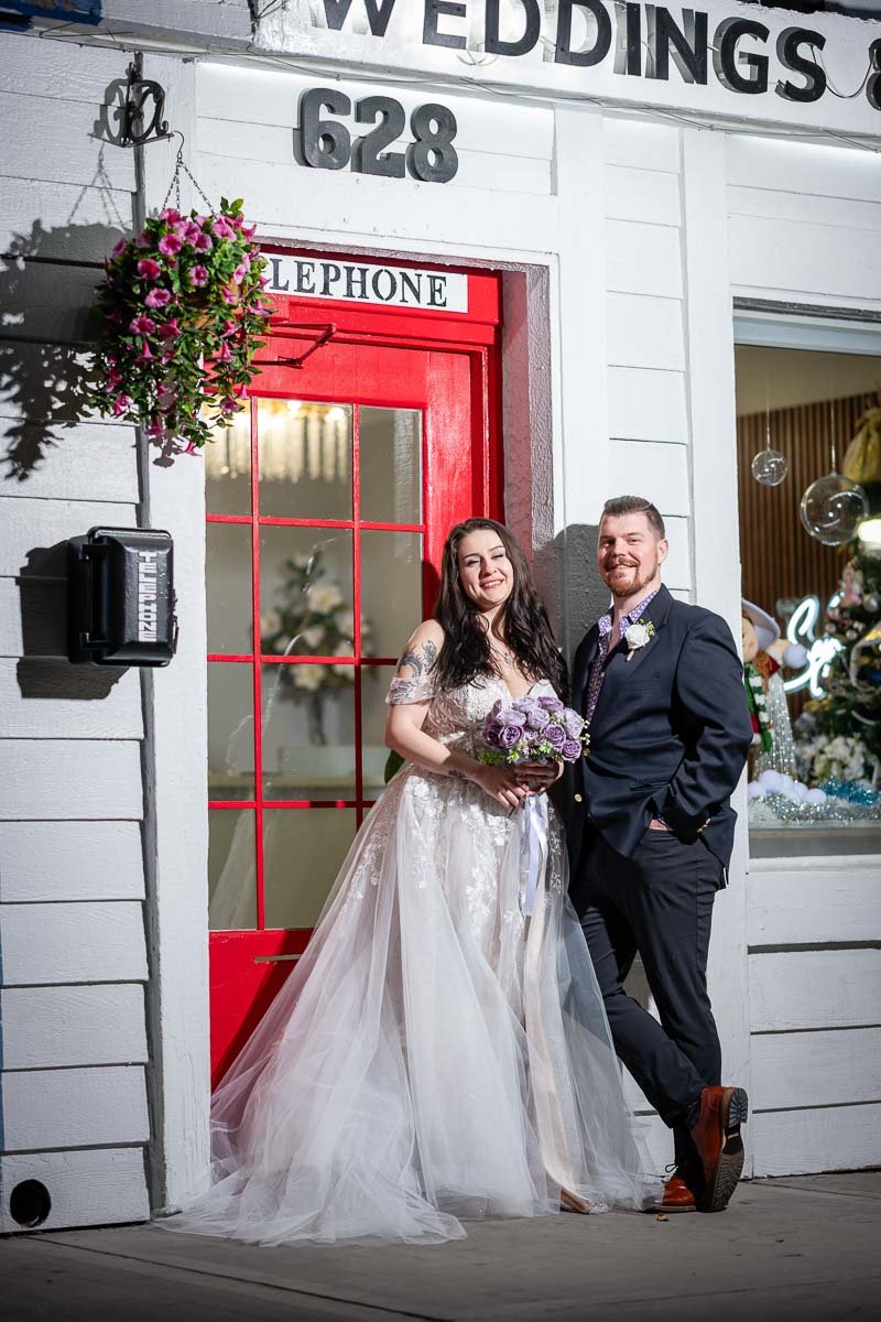A bride and groom standing outside a building with a red door, smiling and posing for a photo, with the bride holding a bouquet of lavender flowers.