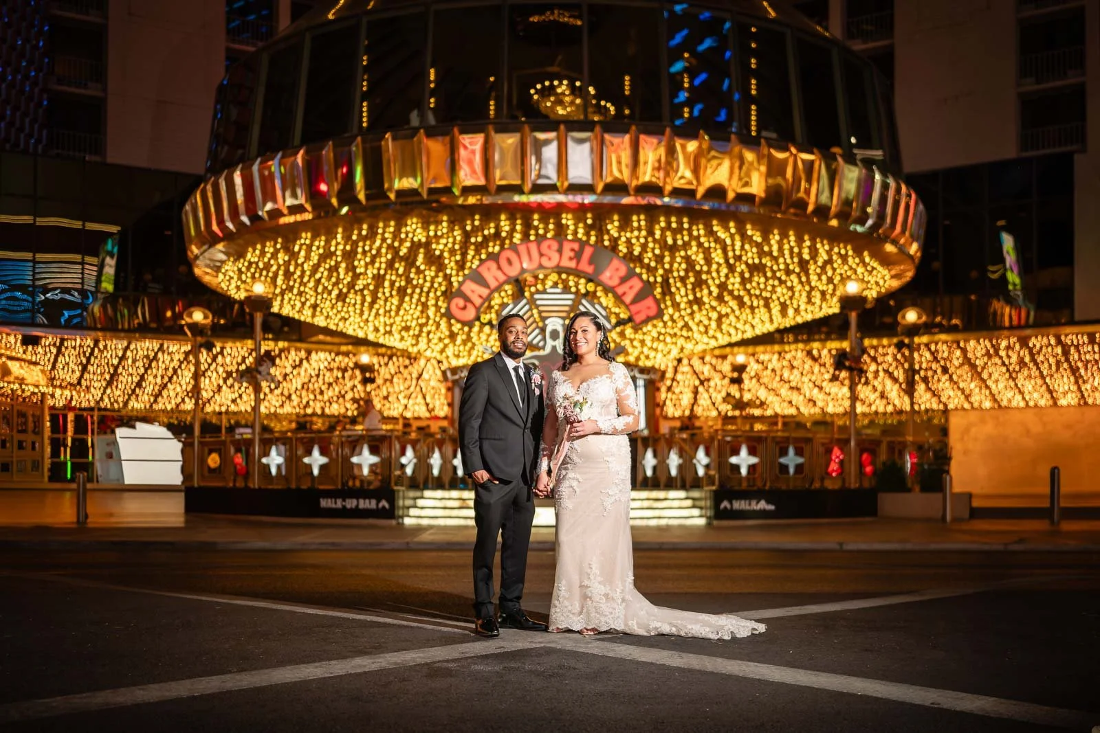 A bride and groom standing in front of a bright, illuminated carousel at night, holding hands and smiling. The carousel has golden lights and a sign that reads 'Carousel Bar'.