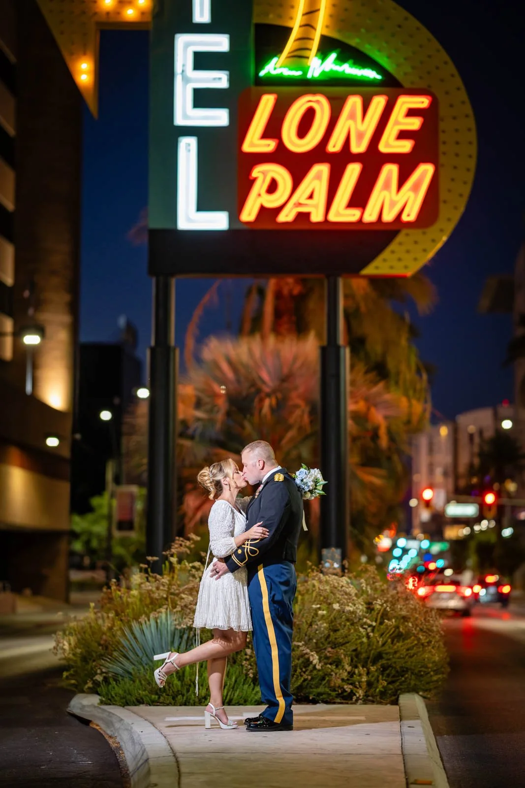 A couple in ceremonial attire sharing an intimate moment at night under the neon sign for the hotel LONE PALM on a city street.