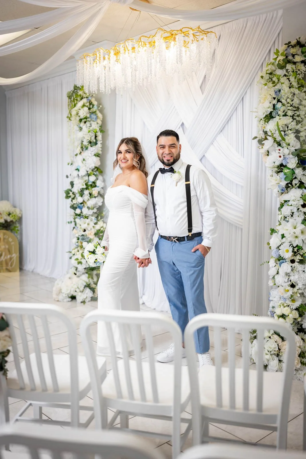 A bride and groom holding hands in a wedding ceremony, standing in front of a floral backdrop with white drapery and a chandelier.