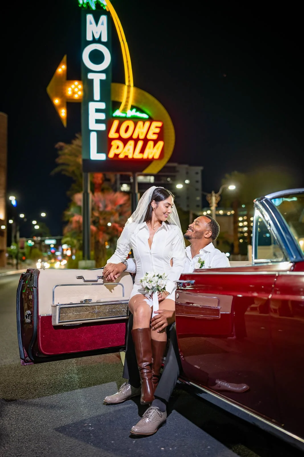A couple in wedding attire sitting on a vintage car at night in front of a neon sign that reads 'Lone Palm Hotel'.
