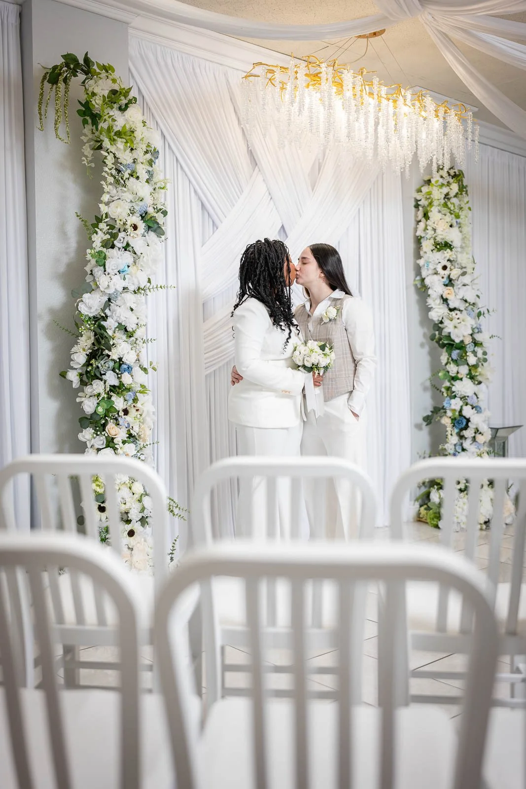 Two women, dressed in white, stand in front of a decorated wedding backdrop, sharing a kiss at the Say I Do Wedding Chapel
