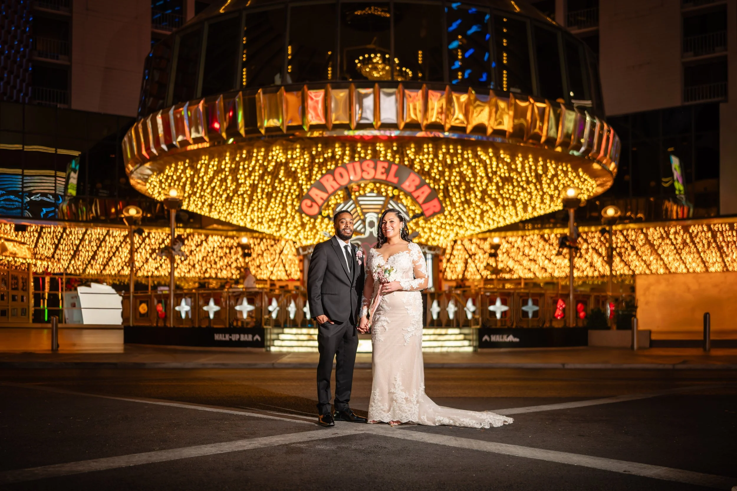 Bride and groom standing in front of a brightly lit carousel at night, holding hands, with the bride in a white lace wedding gown and holding a bouquet, and the groom in a black suit with a tie.
