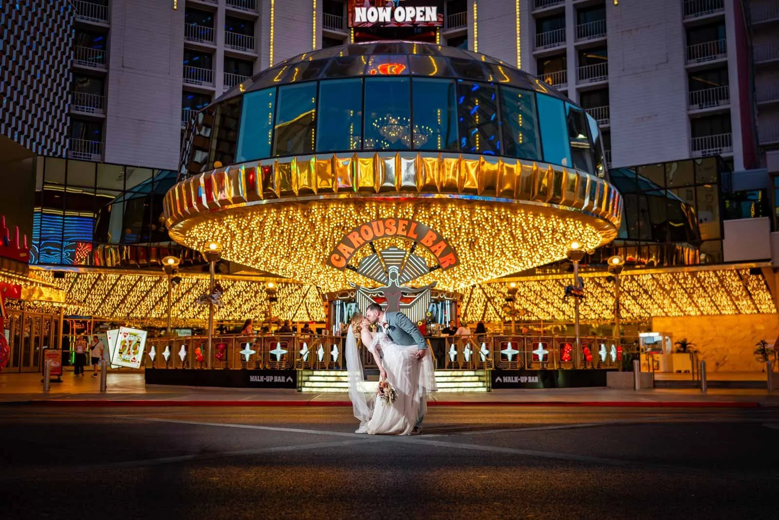 A newlywed couple sharing a dance in front of a brightly lit carousel at night.