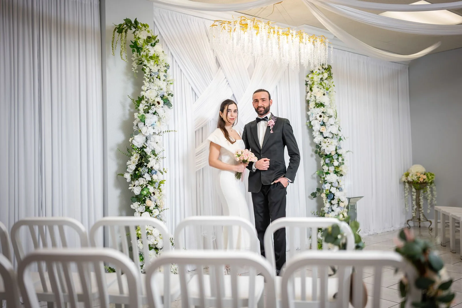 A bride and groom standing together at their wedding ceremony in front of a decorated backdrop of white flowers and draped fabric inside the Say I Do Wedding Chapel in Las Vegas.