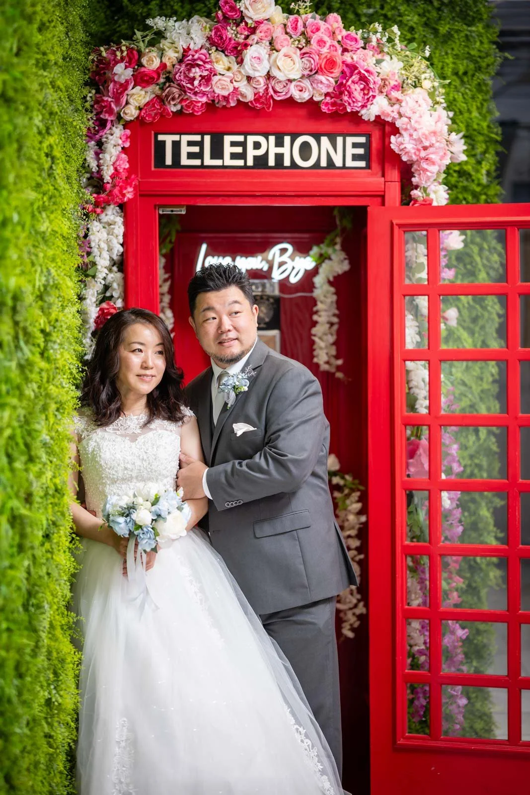 A bride and groom standing in front of a red phone booth decorated with pink and white flowers.