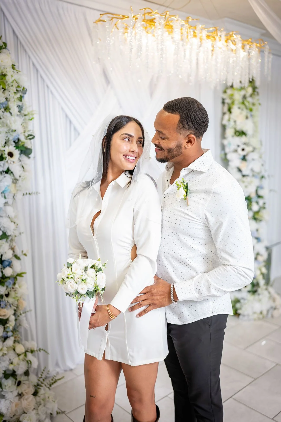 A couple in wedding attire standing close together, smiling at each other, in front of a floral and gold-decorated backdrop at their wedding.
