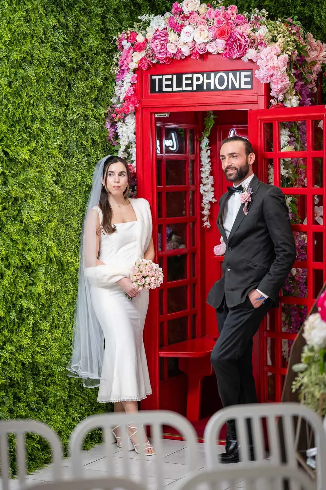A bride and groom pose in front of a red telephone booth inside the Say I Do Cahpel decorated with pink and white flowers at a wedding.