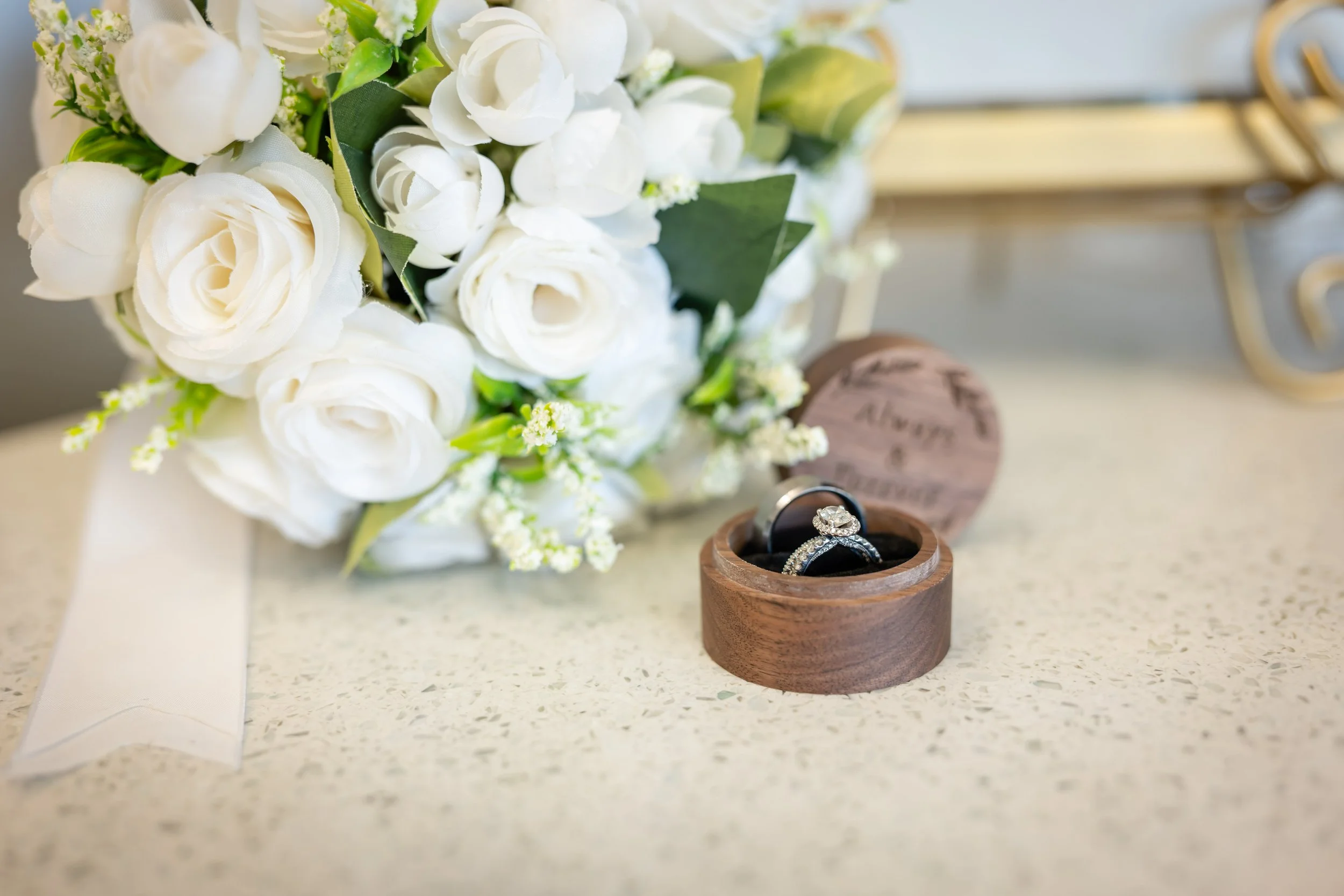 A bouquet of white flowers and a ring inside a wooden box on a countertop, with a small wooden heart-shaped item nearby.