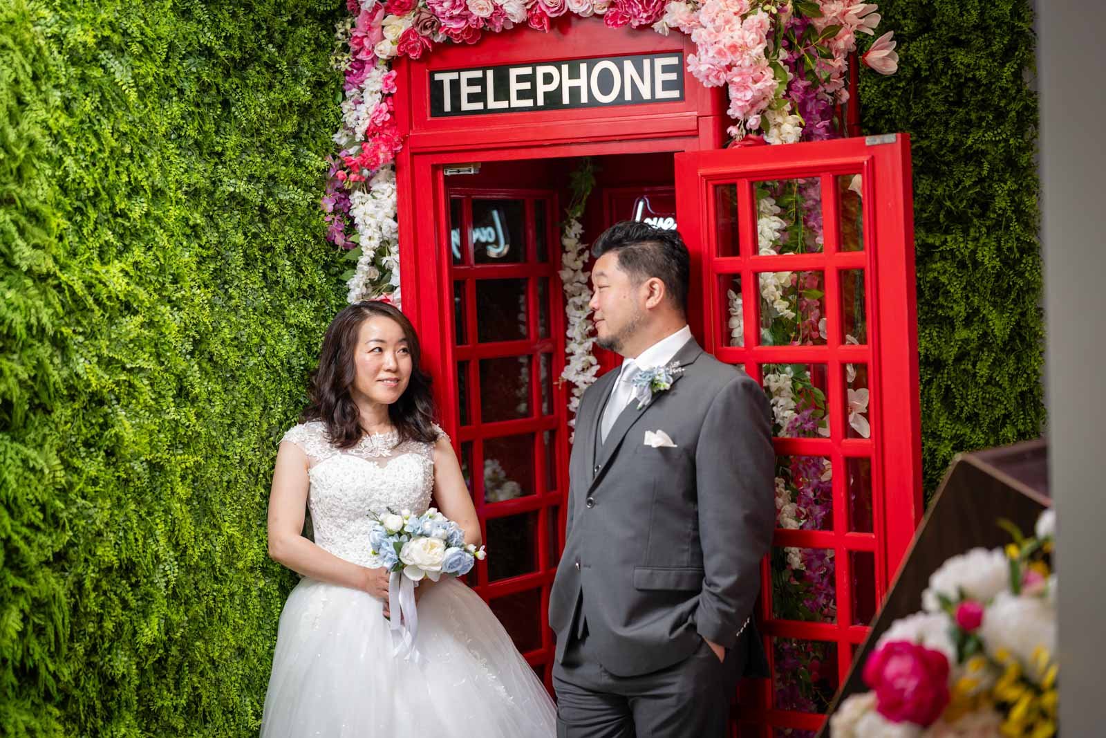 A couple dressed in wedding attire, standing in front of a red telephone booth decorated with flowers, on a lush green floral wall.