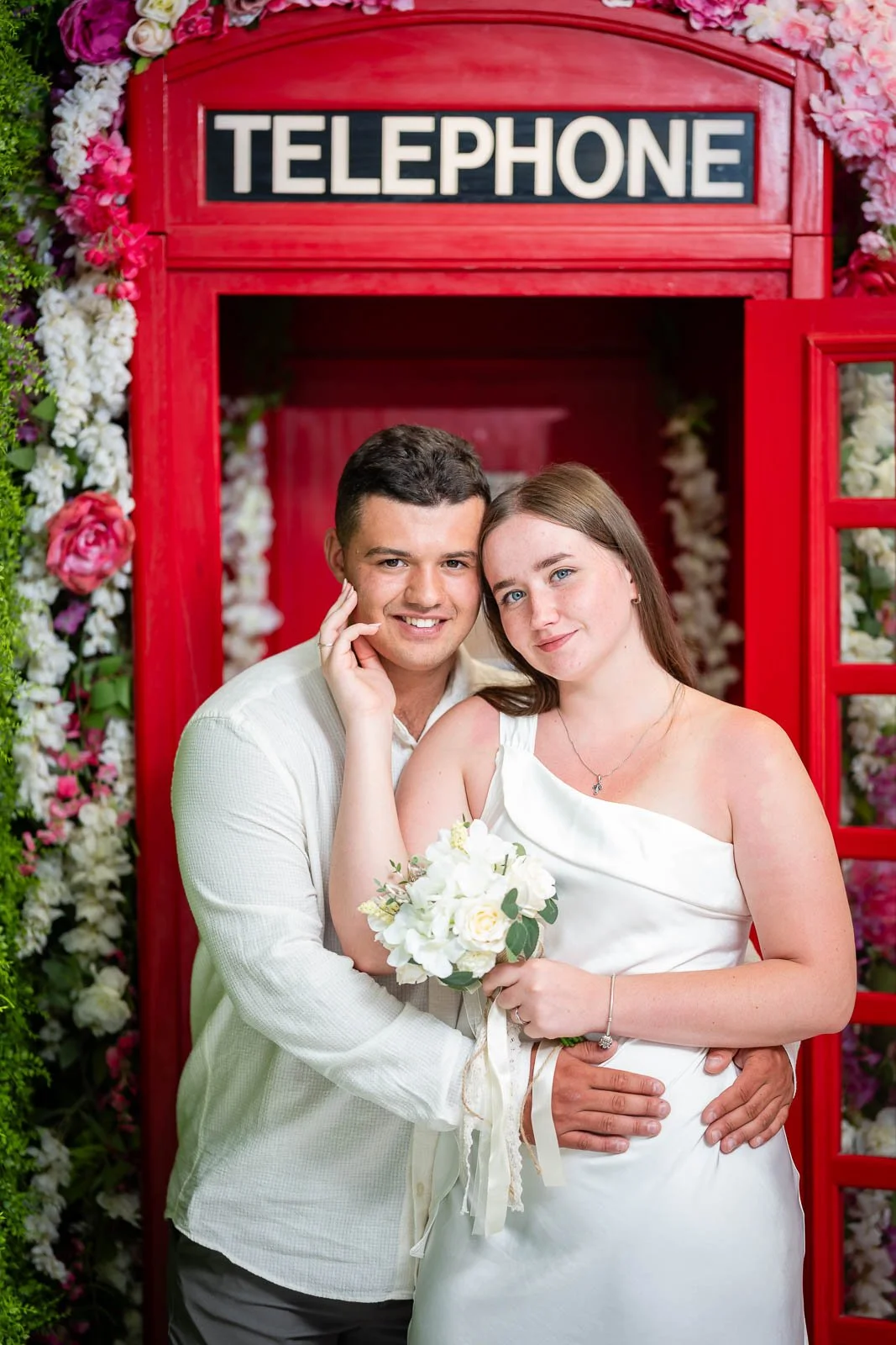 A couple in wedding attire standing in front of a red telephone booth decorated with pink and white flowers, holding a bouquet of white flowers.