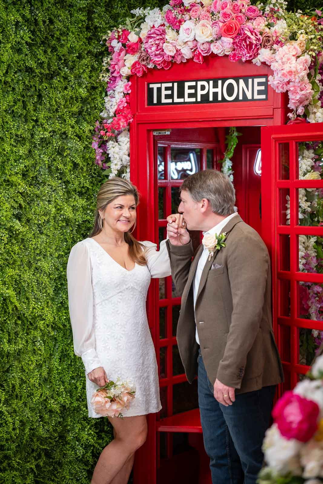 A bride and groom exhibit a playful moment during their wedding, with the groom pretending to kiss the bride's hand inside a romantic, flower-adorned red telephone booth.