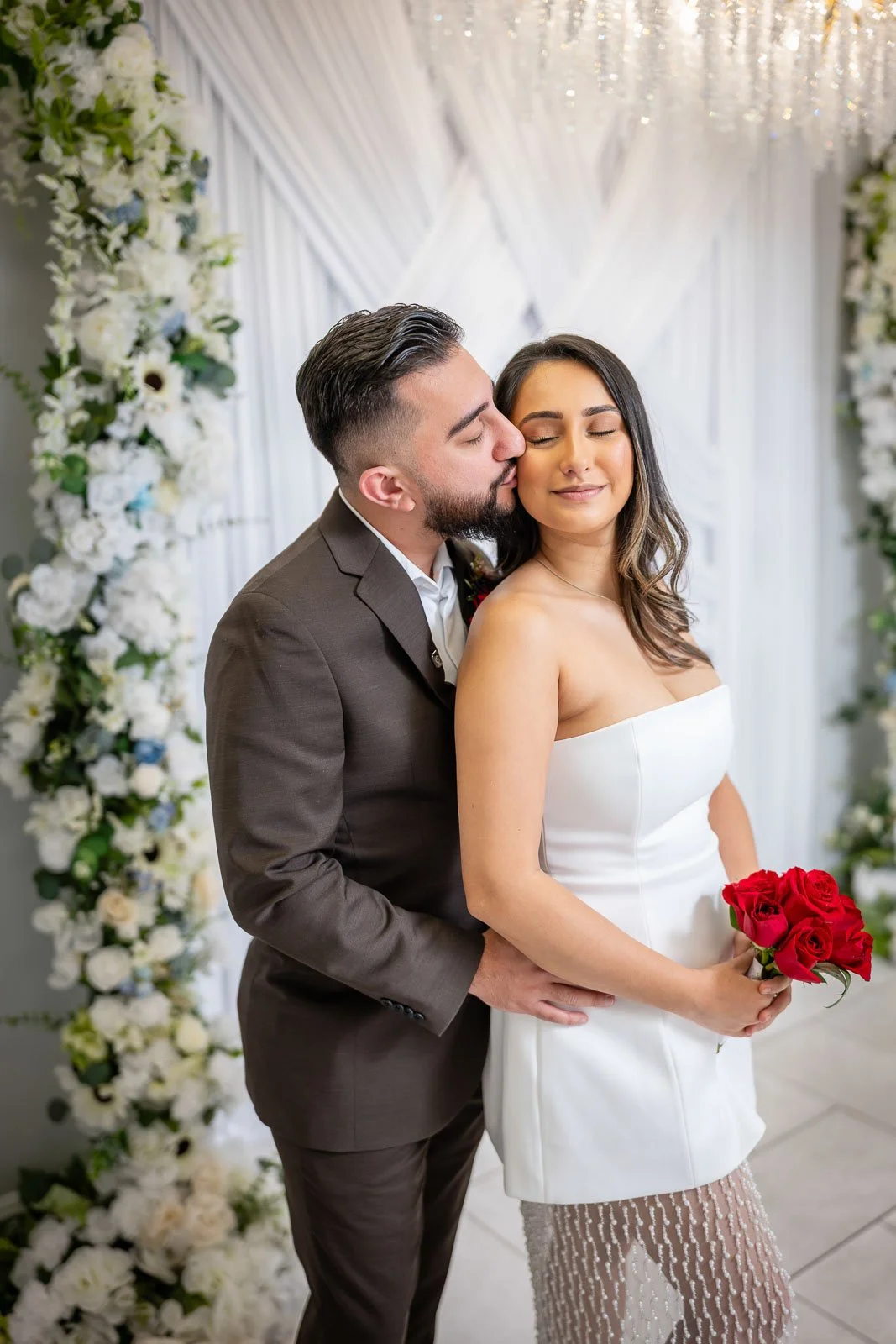 A bride and groom standing together, with the groom kissing the bride's cheek. The bride is holding a bouquet of red roses, and they are in a decorated venue with white floral arrangements and drapes in the background.