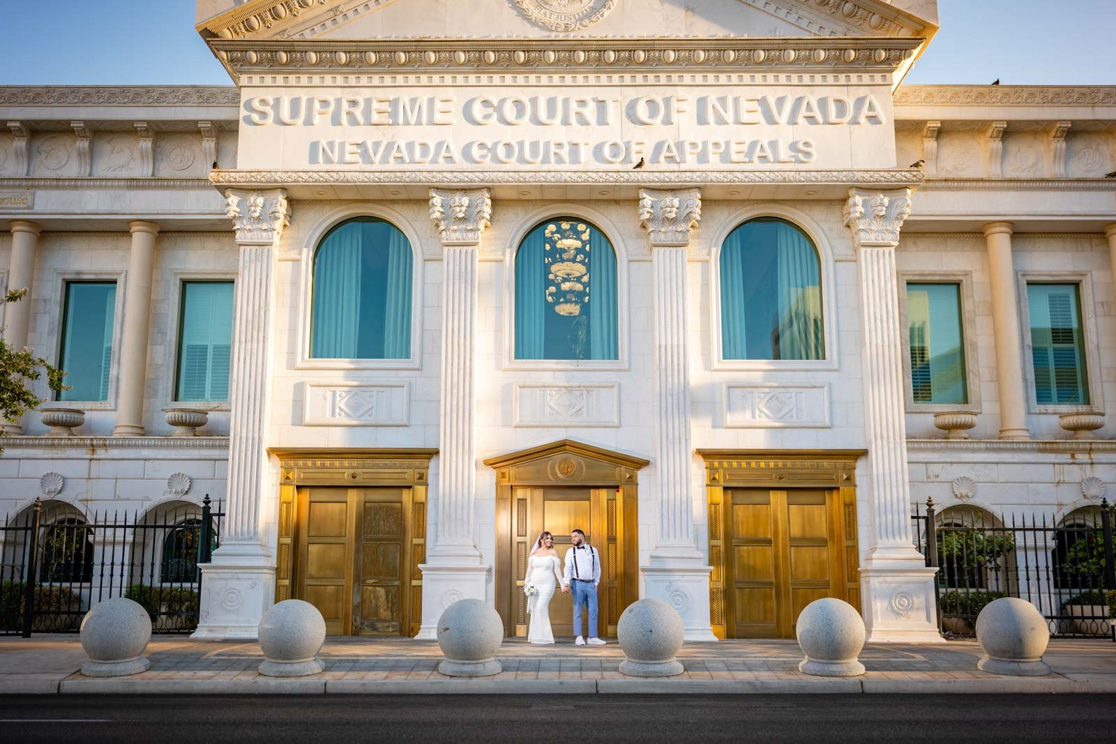A couple dressed in wedding attire standing hand in hand in front of the Supreme Court of Nevada building. The building has a white facade with tall columns and gold accents, with large arched windows and the courthouse name inscribed at the top.