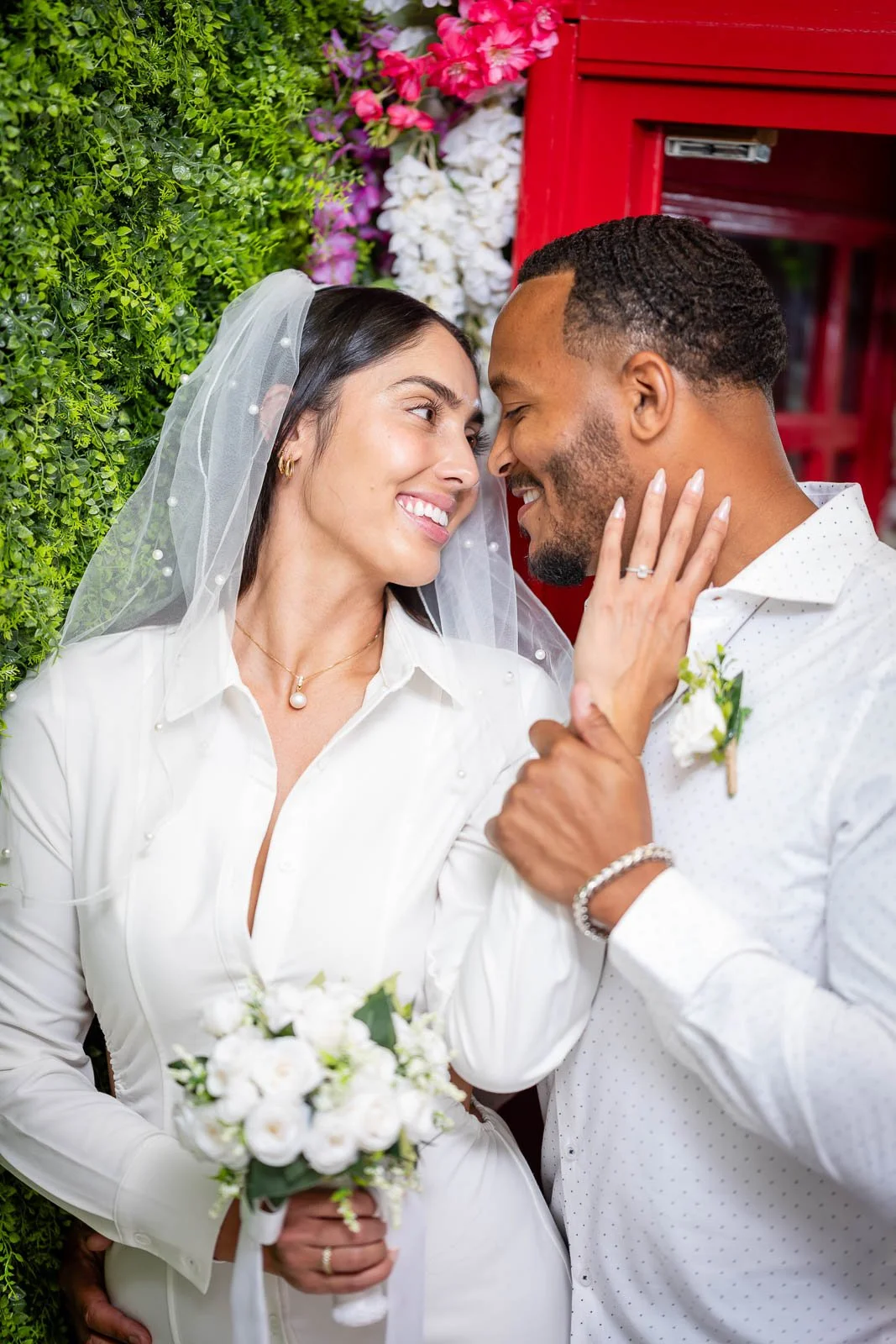 A newlywed couple sharing an intimate moment in front of a green hedge and pink, white, and purple flowers. The bride is holding a bouquet of white roses and wearing a white dress with a veil, while the groom is dressed in a white shirt with a bouton