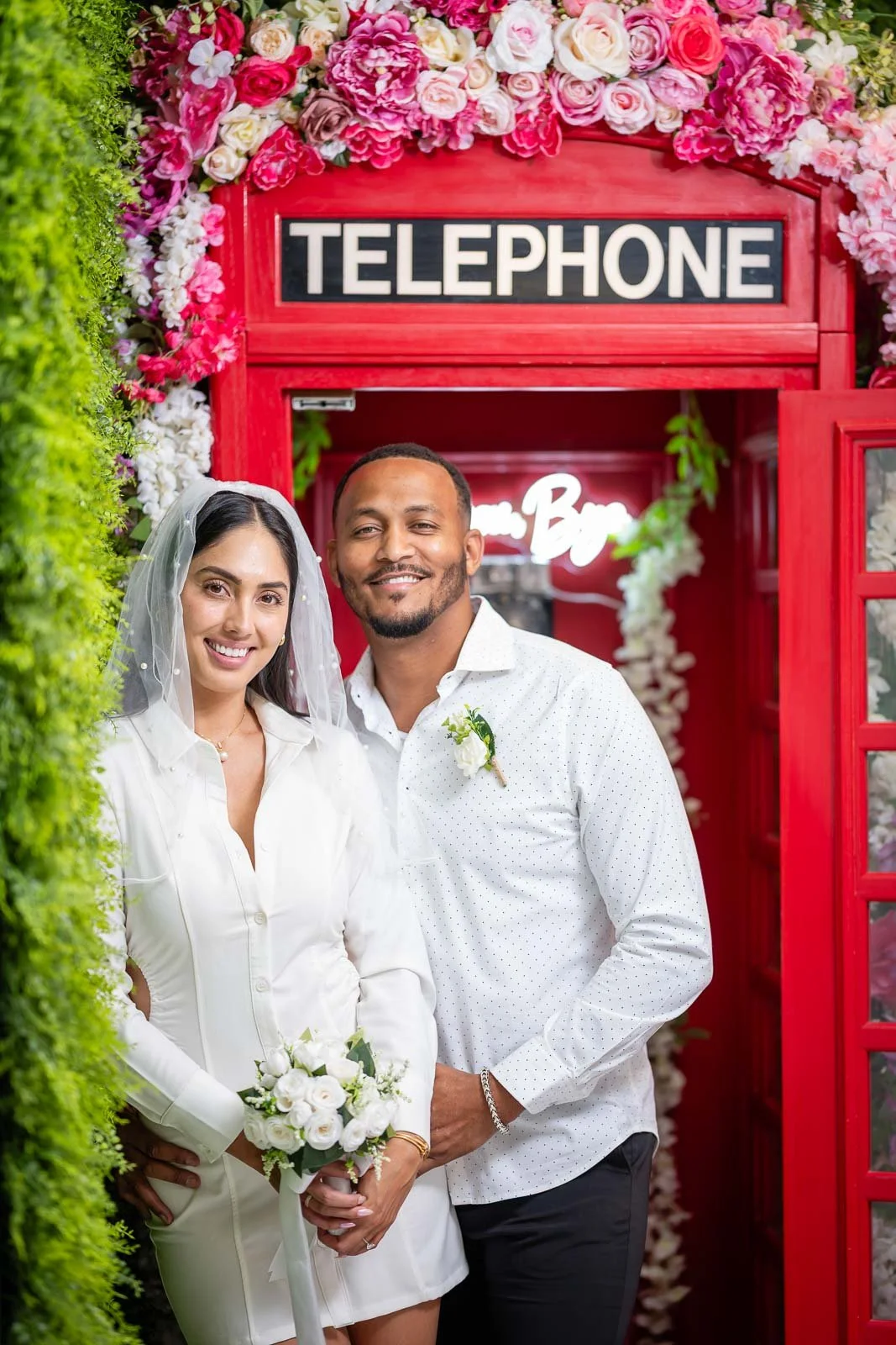 A bride and groom smiling and standing close together, holding a bouquet. They are in front of a decorated red telephone booth adorned with pink and white flowers, with a vivid green hedge on one side.
