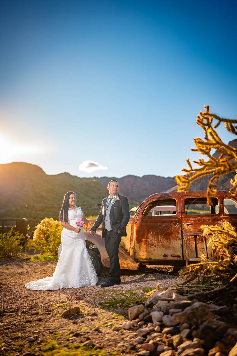 A bride and groom in wedding attire holding hands next to a rusted vintage car in a desert landscape at sunset.