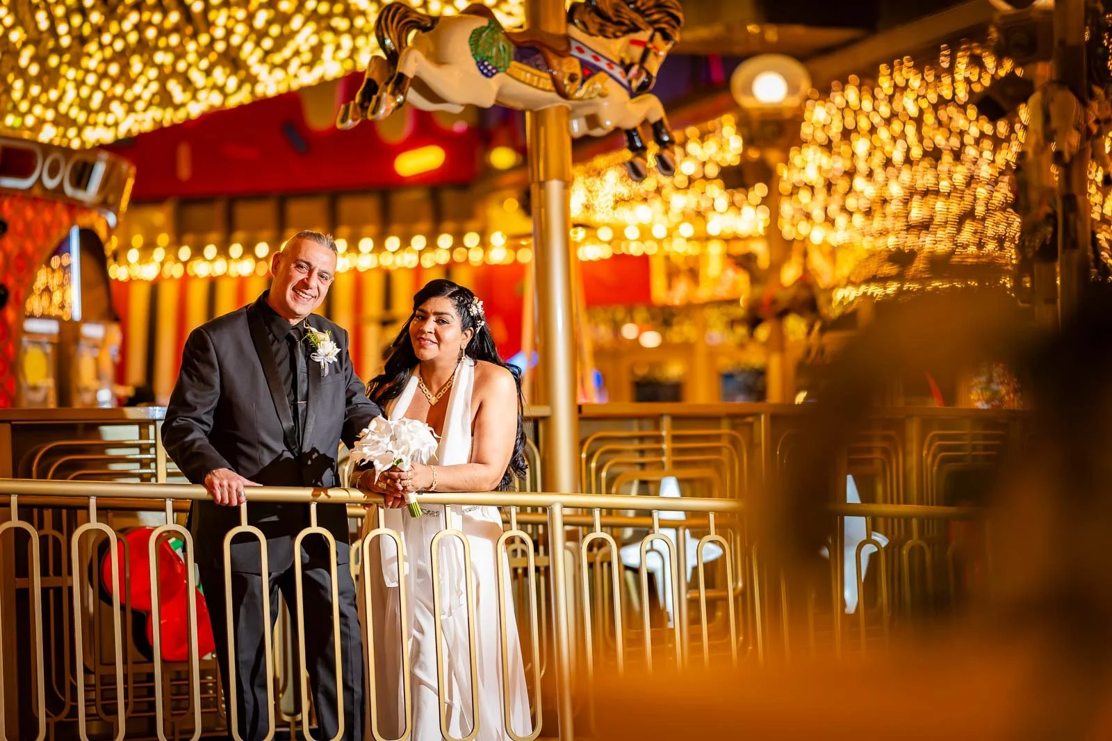 A newlywed couple standing on a carousel at night, surrounded by bright, warm-colored lights. The groom is wearing a black suit with a boutonnière, and the bride is wearing a white dress holding a bouquet, both smiling.