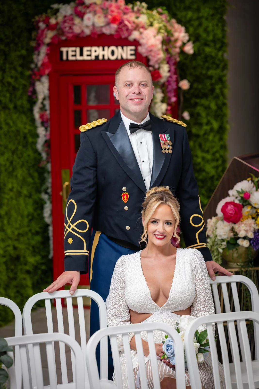A man in a military dress uniform and a woman in a white dress sitting at a wedding ceremony with flowers and a red telephone booth in the background.