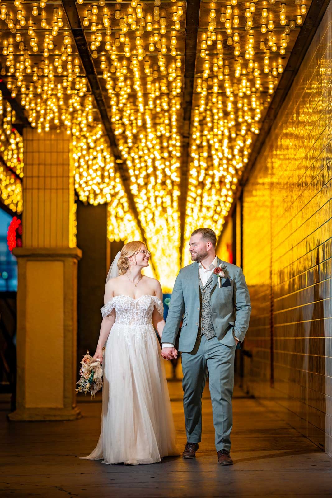 A couple in wedding attire walking hand in hand under a canopy of warm yellow string lights at night.