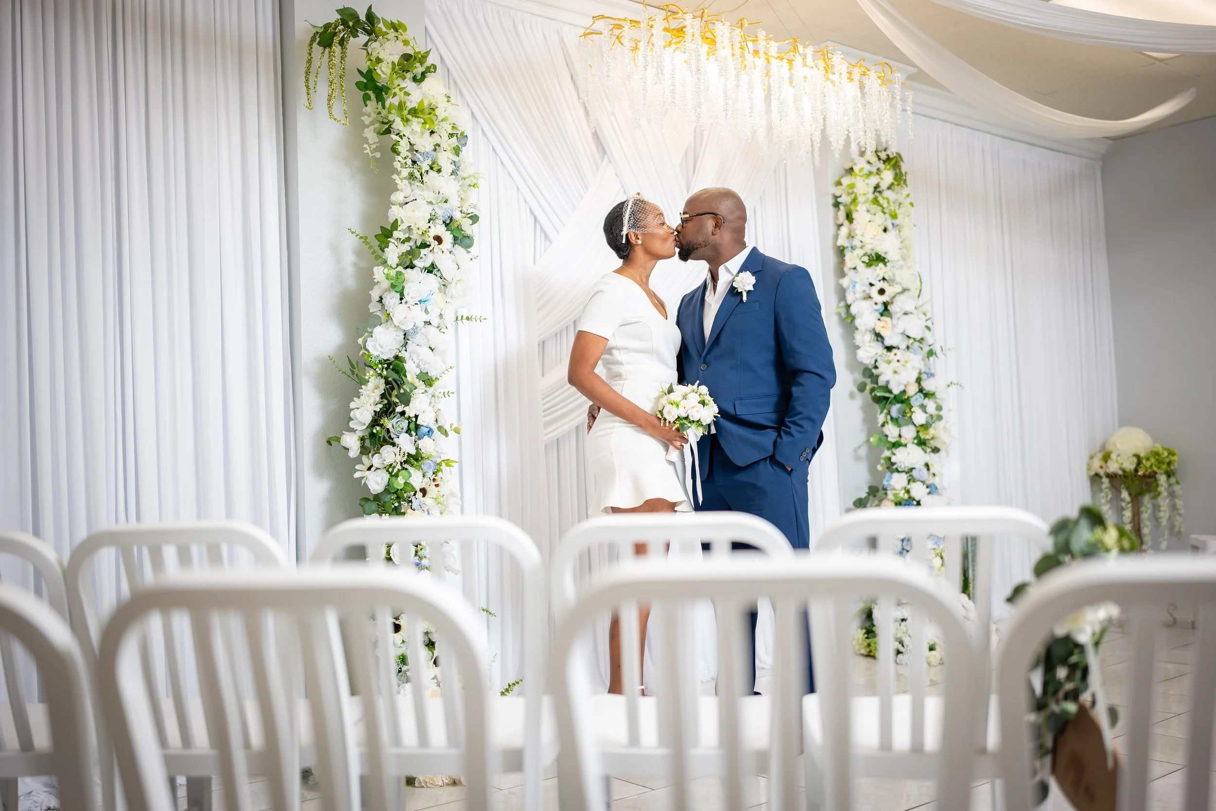 Couple kissing at their wedding ceremony in a decorated white venue with floral arch and chairs.