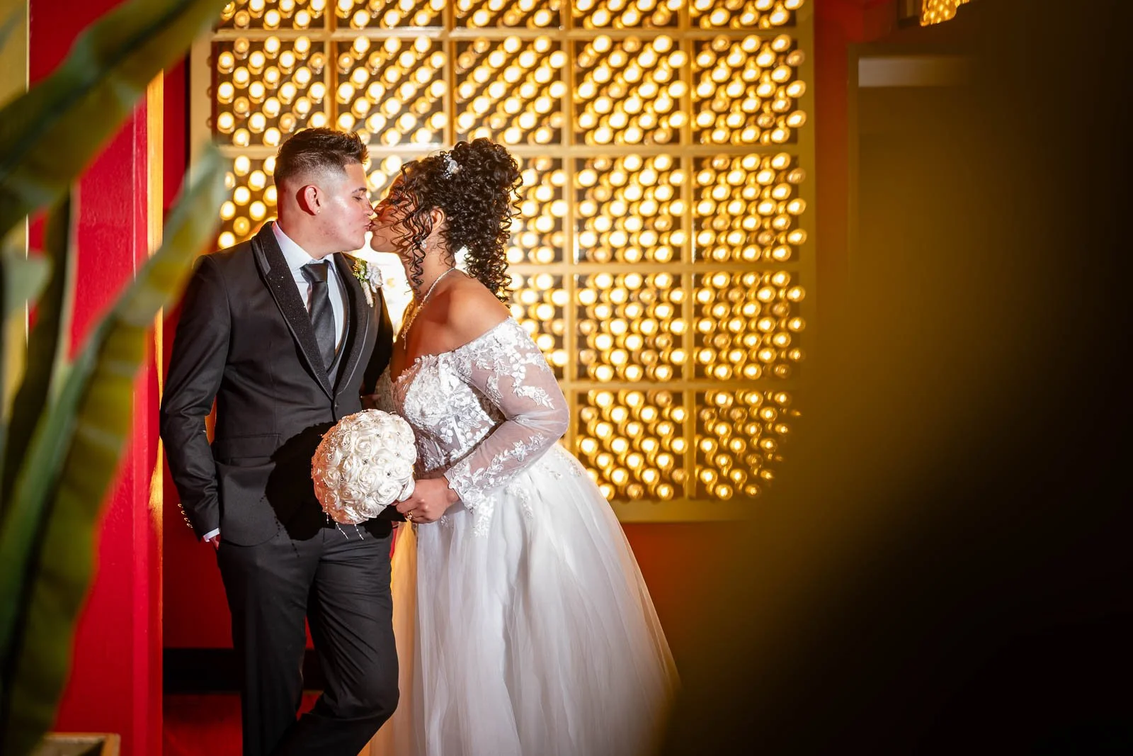 A newlywed couple sharing a kiss indoors, with the bride holding a bouquet of white roses and wearing a white off-shoulder wedding dress with lace details, and the groom in a black suit with a black tie, against a background of warm glowing lights.