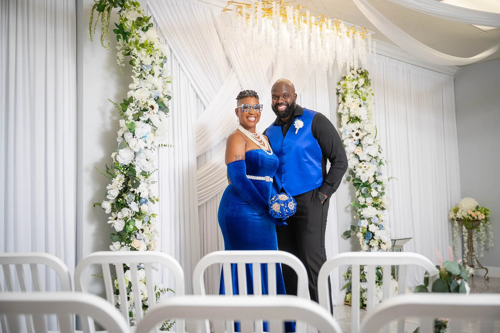 A smiling couple dressed in blue and black standing in front of a white, floral wedding backdrop.
