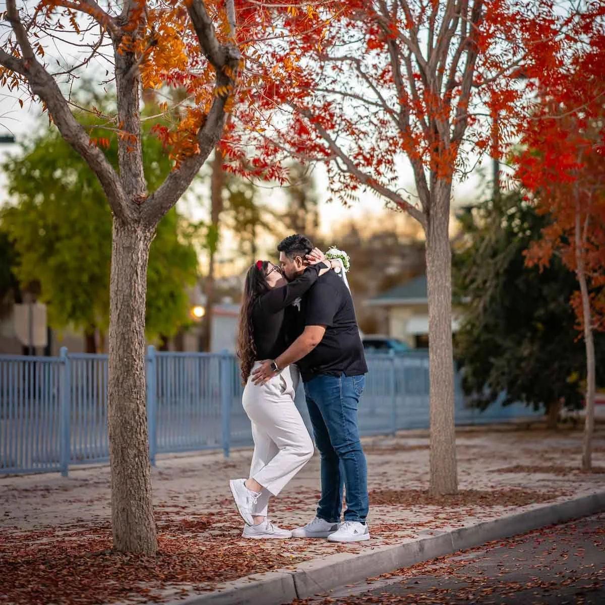 A couple sharing a romantic moment under orange autumn trees in an outdoor park setting during sunset.