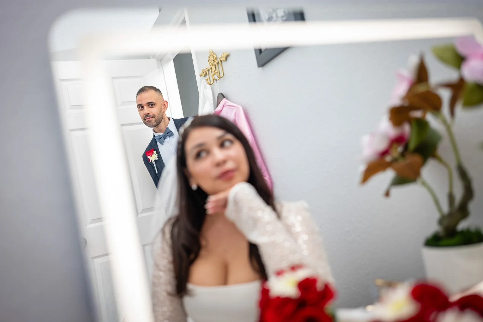 A bride with dark hair looking at a man in a suit reflected in a mirror, with pink flowers and a white door in the background.