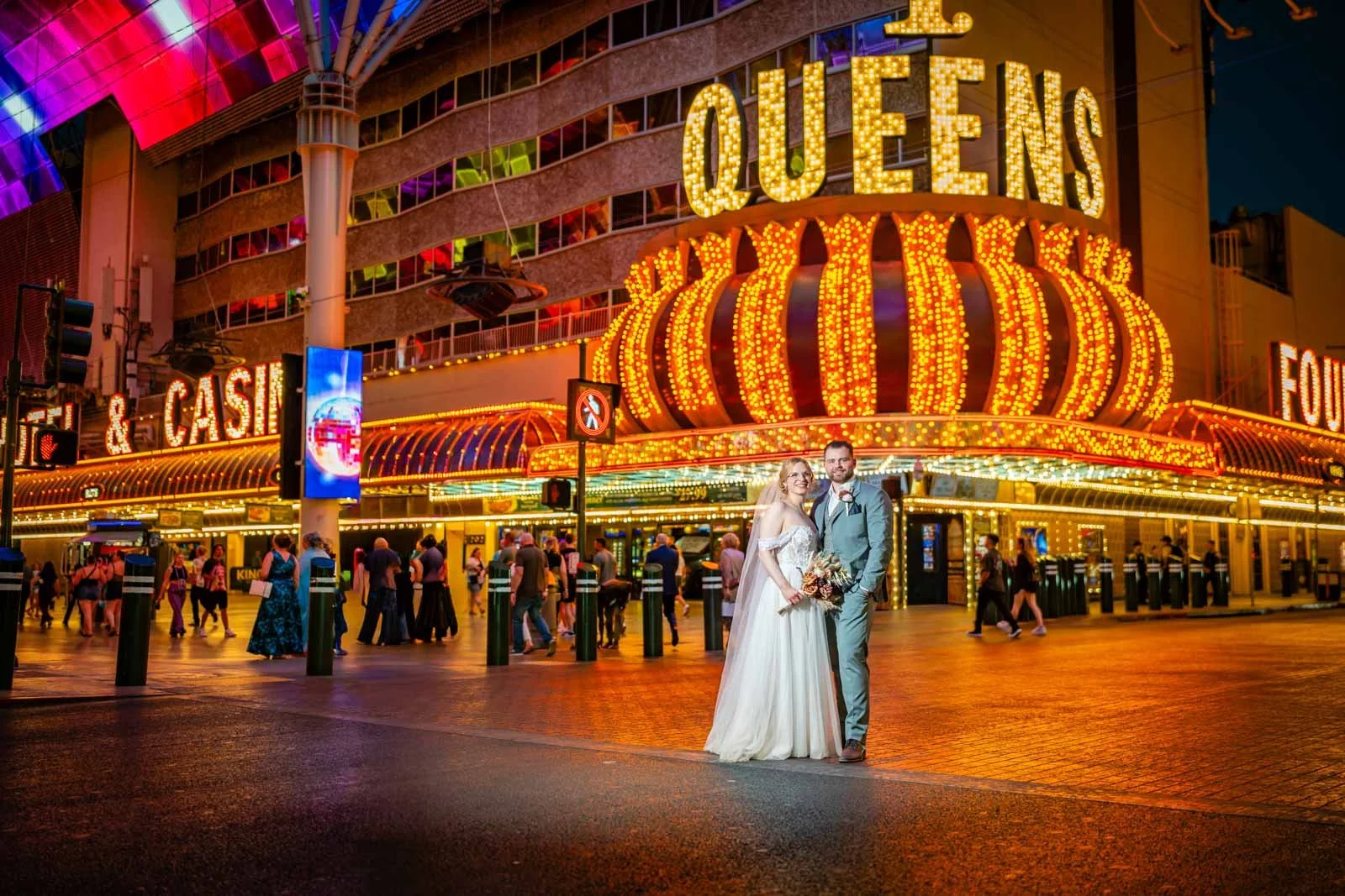A couple in wedding attire standing in front of the illuminated Queen's Theater in Las Vegas at night, with a crowd of people walking nearby and vibrant neon lights