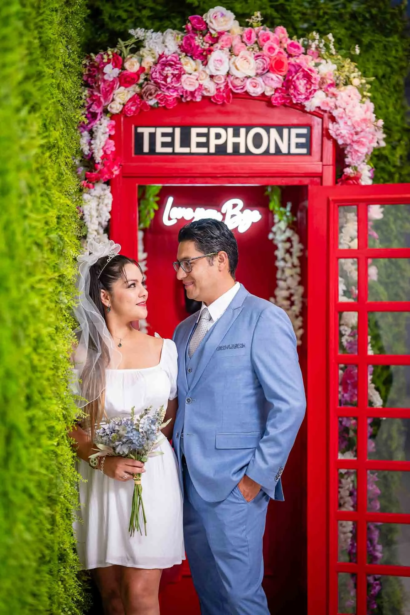 Couple dressed in wedding attire standing close together in front of a bright red telephone booth decorated with pink and white flowers, with a green foliage wall on the left side.