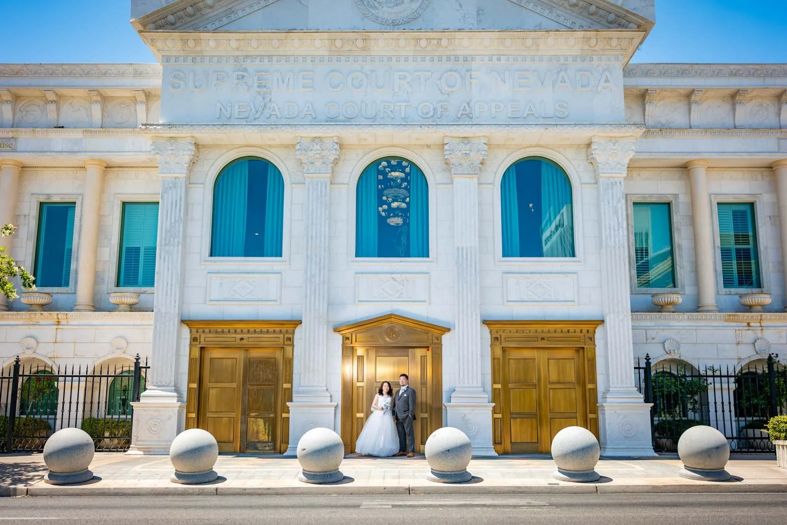 A bride and groom standing in front of the Supreme Court of Nevada, a white building with large columns and golden doors, with reflections of the street and blue curtains in the windows.