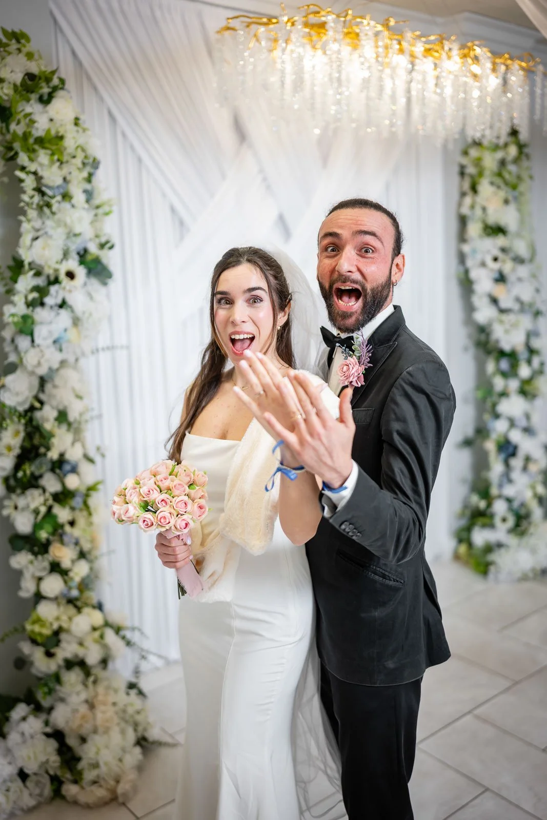 A bride and groom celebrating their wedding, smiling and showing their wedding rings, with floral decorations and a chandelier in the background.