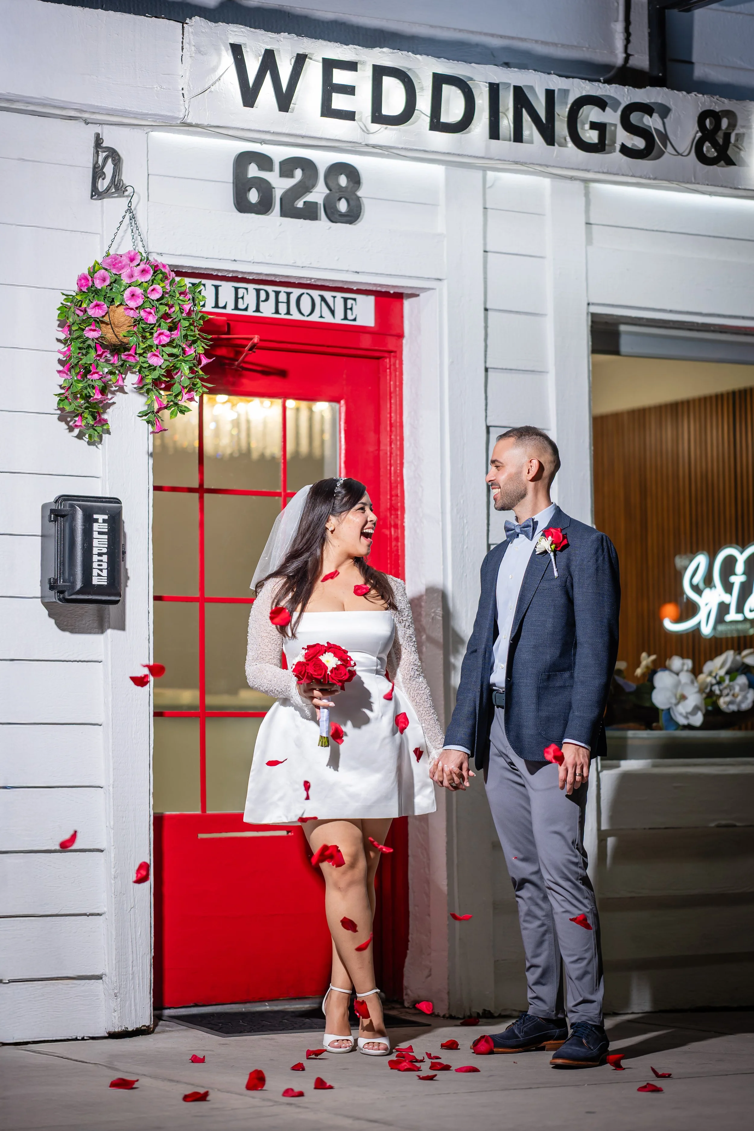 A newlywed couple standing outside a venue with a sign above that reads 'WEDDINGS & 628'. The bride is wearing a white dress with a veil, holding a bouquet, and smiling at the groom. The groom is dressed in a blue suit with a bowtie, smiling at the b