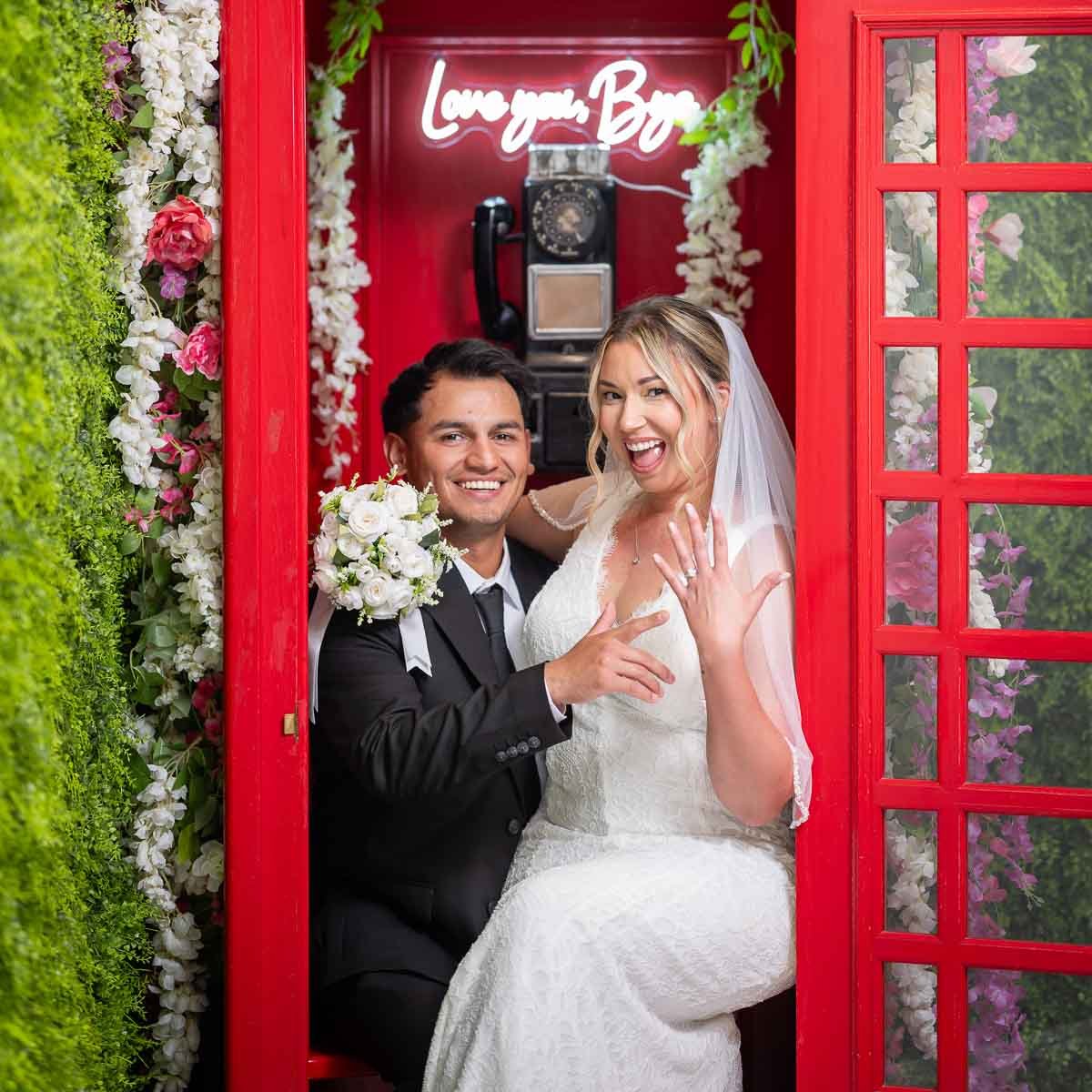 A happy bride and groom inside a red phone booth decorated with flowers, with a neon sign that says 'Love you Bae' and a vintage phone inside.