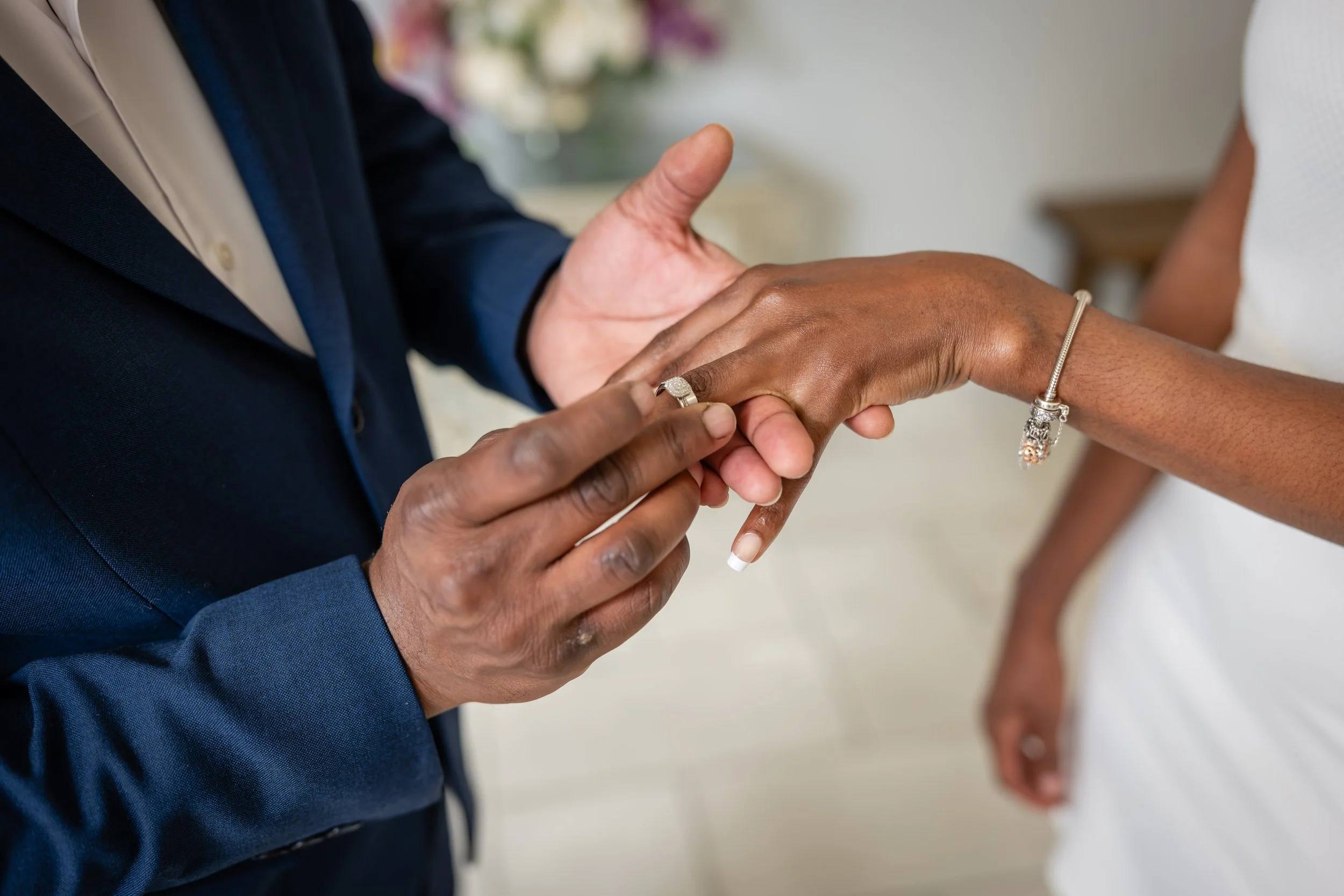 A person in a dark suit is placing a ring on the finger of another person wearing a white dress, during a wedding ceremony.