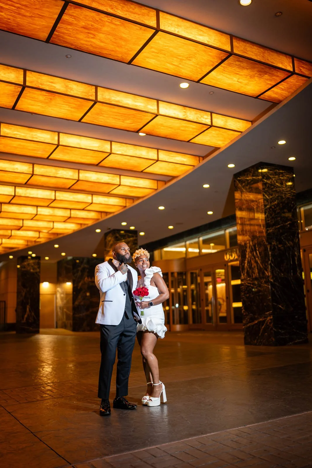 A happy couple posing for a photo under a warm, glowing ceiling in a spacious, elegant lobby. The man is dressed in a tuxedo, and the woman is wearing a white dress with high heels, holding a bouquet of red roses.