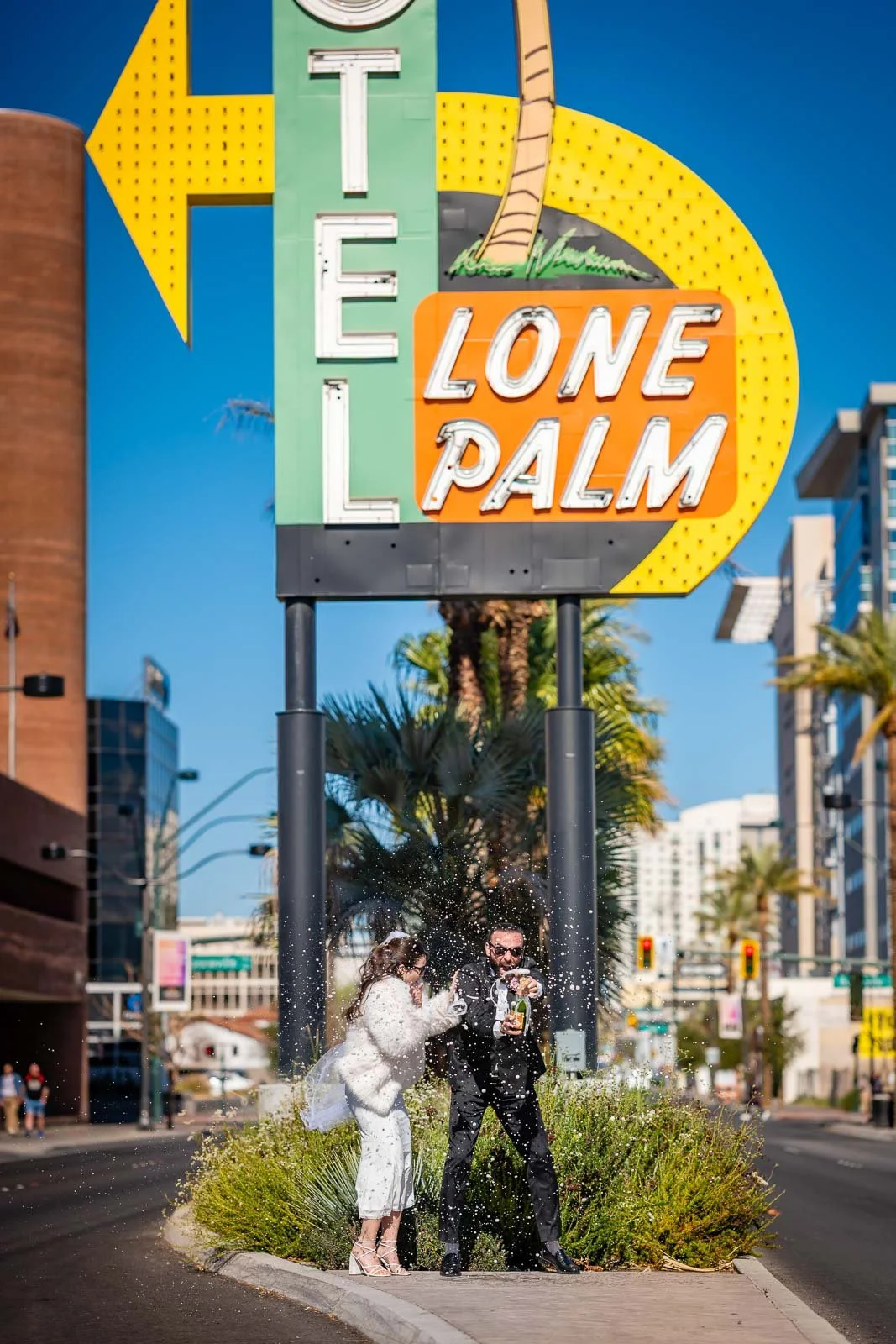 Couple in wedding attire celebrating under the iconic Lone Palm sign in Las Vegas, with water splashing near their feet and a cityscape in the background.