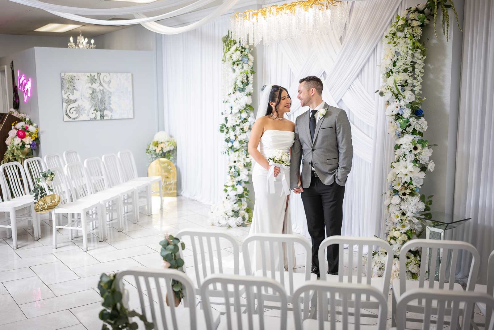 Bride and groom smiling at each other during their wedding ceremony in a decorated indoor venue with white chairs and floral arrangements.