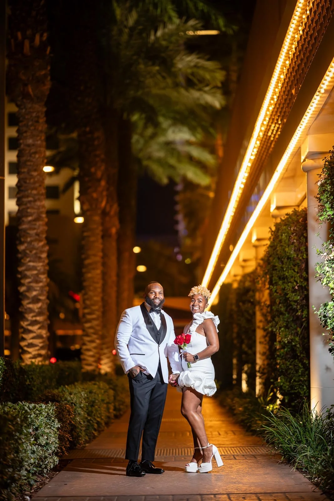 A couple dressed in formal attire, standing outdoors on a night illuminated walkway, with palm trees and decorative lighting behind them.