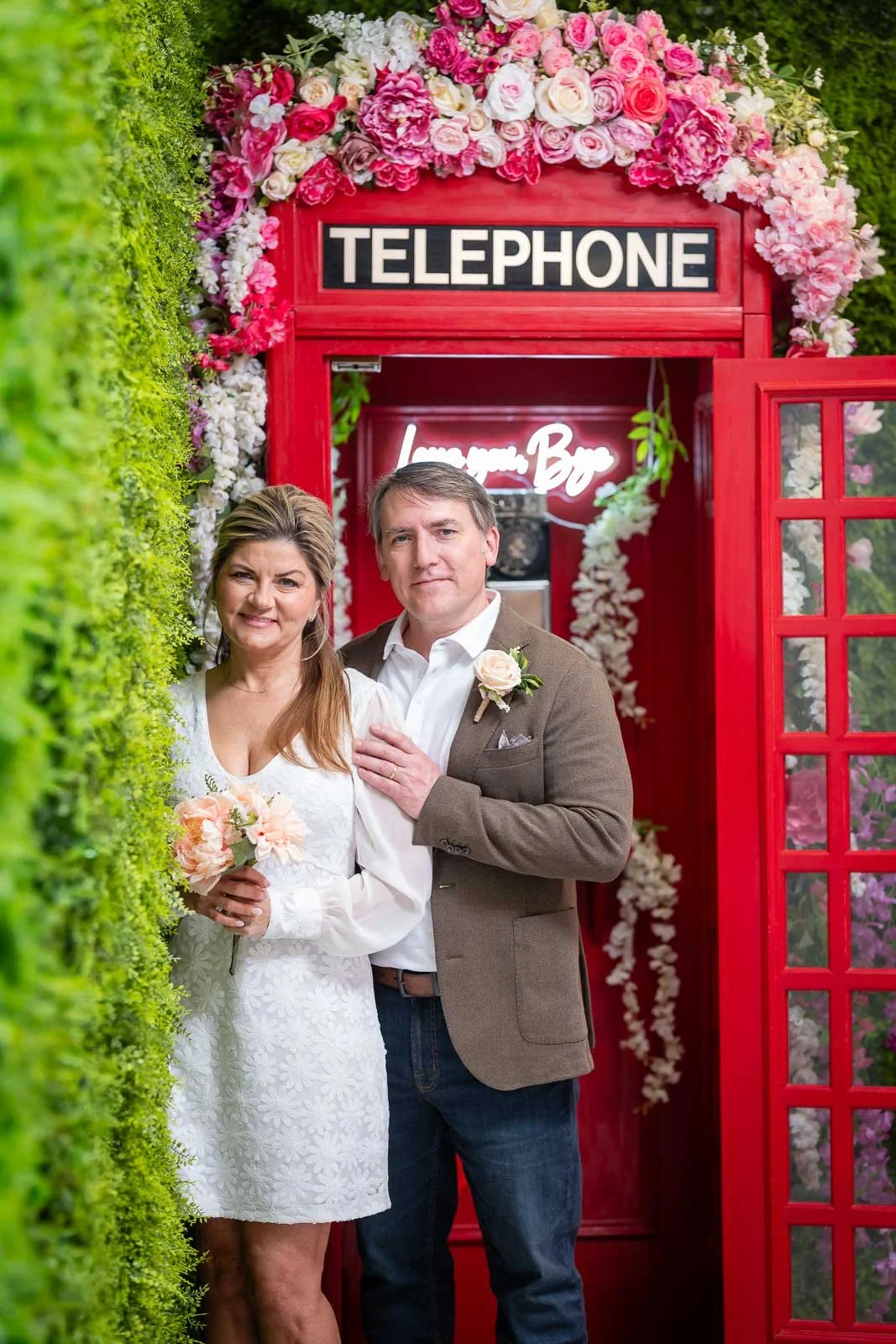 A middle-aged couple standing inside a decorative red telephone booth, surrounded by pink and white flowers, with a neon sign that says 'Love you, Bar' in the background.