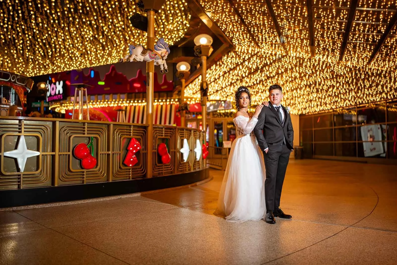 A bride and groom standing together at a night amusement park decorated with bright yellow string lights, with the bride in a white wedding dress and the groom in a black tuxedo.