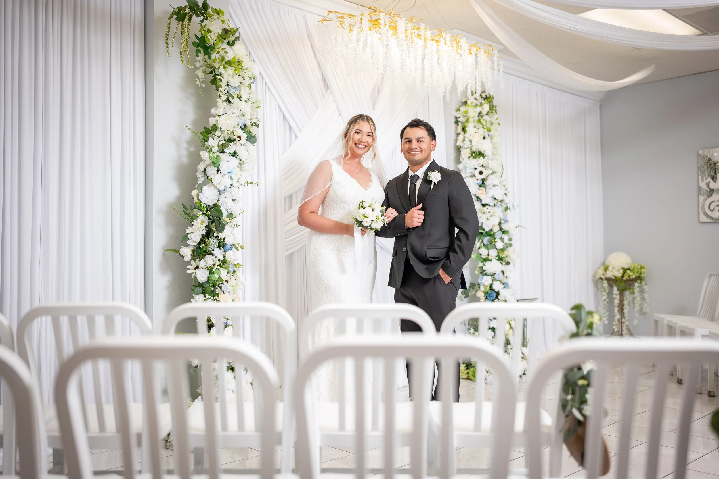 A bride and groom standing side by side, smiling, in front of a floral wedding arch. The bride is wearing a white lace wedding dress and holding a bouquet, while the groom is in a black suit with a white shirt and black tie. The background is decorat
