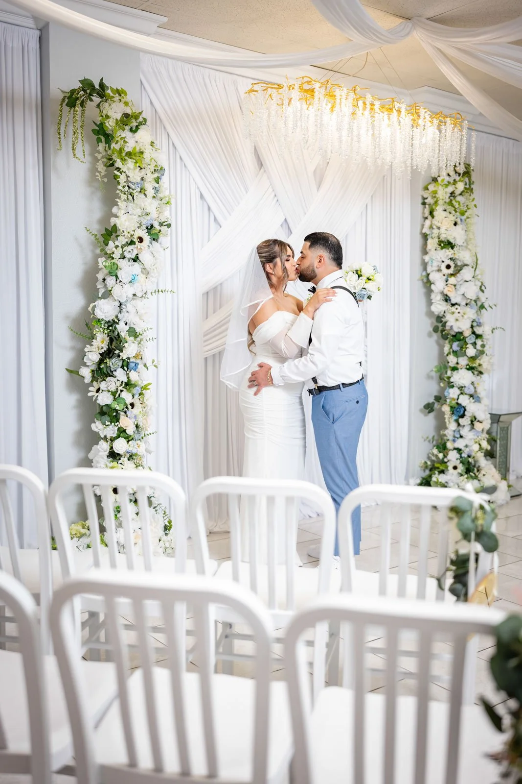 A bride and groom sharing a kiss during their wedding ceremony under a white floral and draped backdrop.