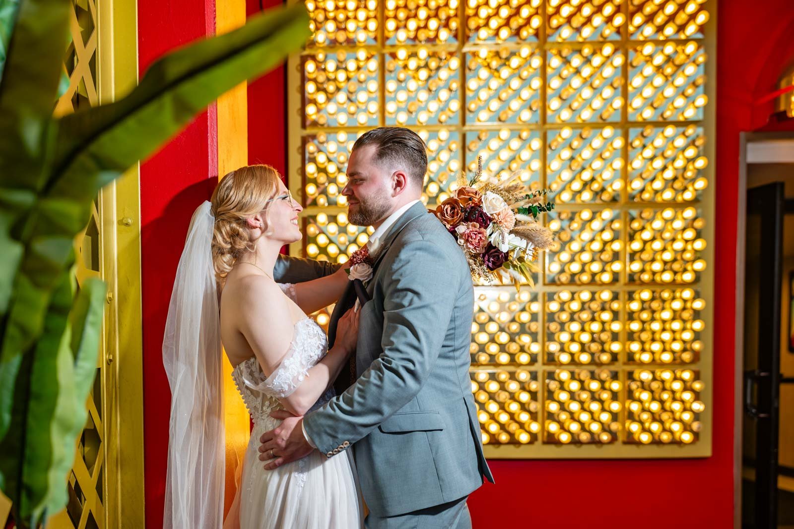 A bride and groom greeting each other indoors, with warm lighting and a colorful decorative background, showing a romantic moment during their wedding celebration.
