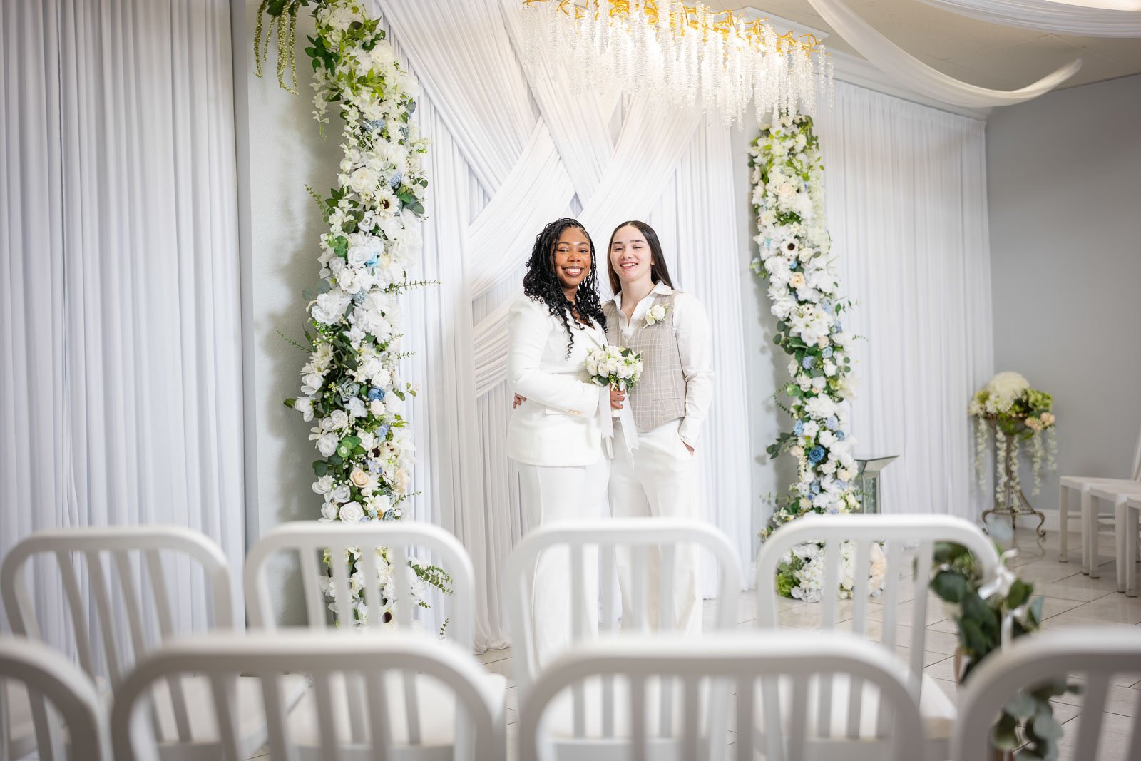 Two women in wedding attire standing under a decorated wedding arch, smiling in the Say I Do Wedding Chapel in Las Vegas