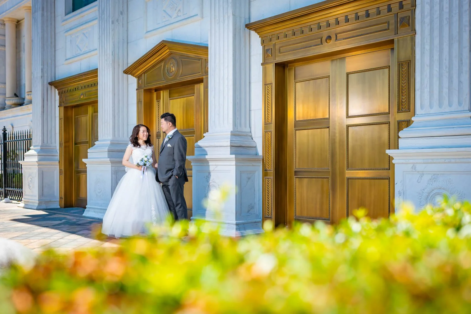 A bride and groom standing outdoors in front of large gold doors with white marble columns, with yellow and green flowers in the foreground.